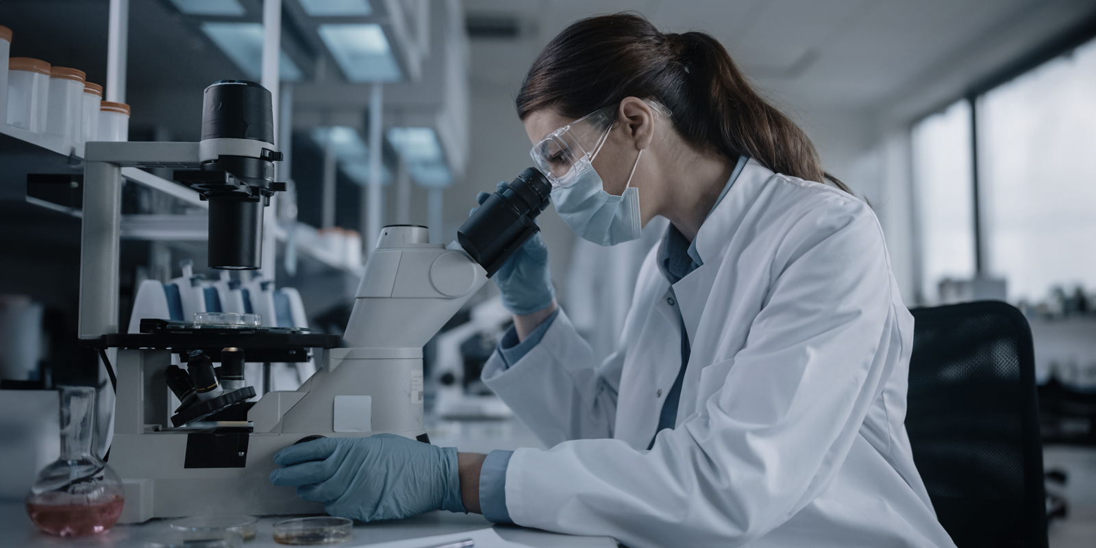 person in lab coat and other PPE look into microscope